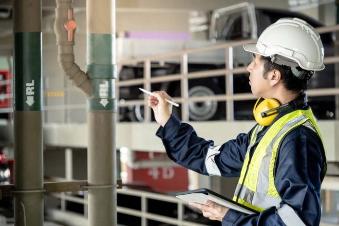 Building inspector man using digital tablet pointing at plumbing pipeline system. Asian male worker in green vest, protective ear muffs and safety helmet working for building maintenance inspection representing Building maintenance backlog and retrospective digitisation