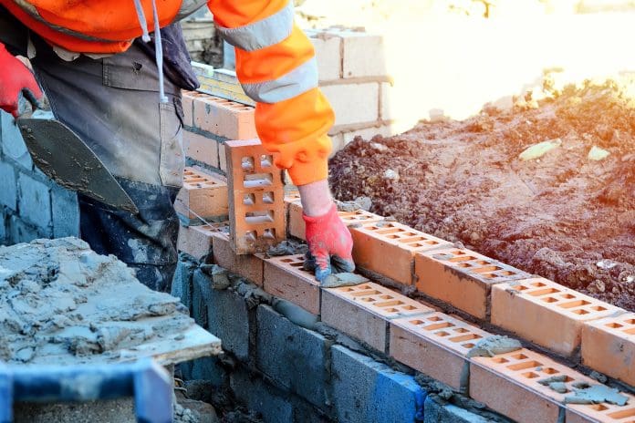 Industrial bricklayer laying bricks on cement mix on construction site. Fighting housing crisis by building more affordable houses concept, representing digital technologies