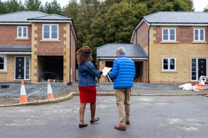 Medium rear-view shot of a female estate agent standing with a male viewer at a housing development site in Northumberland, North East England. The estate agent is holding paper plans as they have a discussion, incomplete houses visible in front of them, representing unfinished projects being brought to market