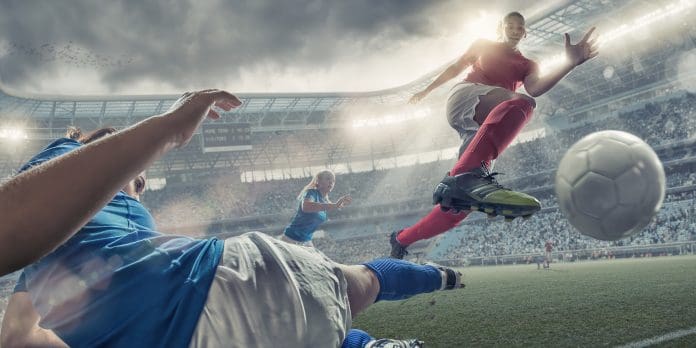 A cross processed mid action image of professional women soccer player in mid air jumping over a rival player who is performing a sliding tackle. The action takes place during a professional women’s football game in a generic floodlit soccer stadium full of spectators under a stormy evening sky at sunset, representing the discussion around women's sports venues