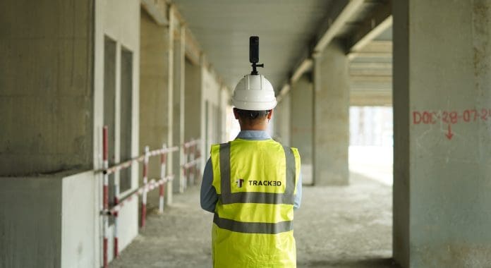 A construction worker stands with back to camera in hi vis and hard helmet mounted with a 3D camera used for AI progress monitoring