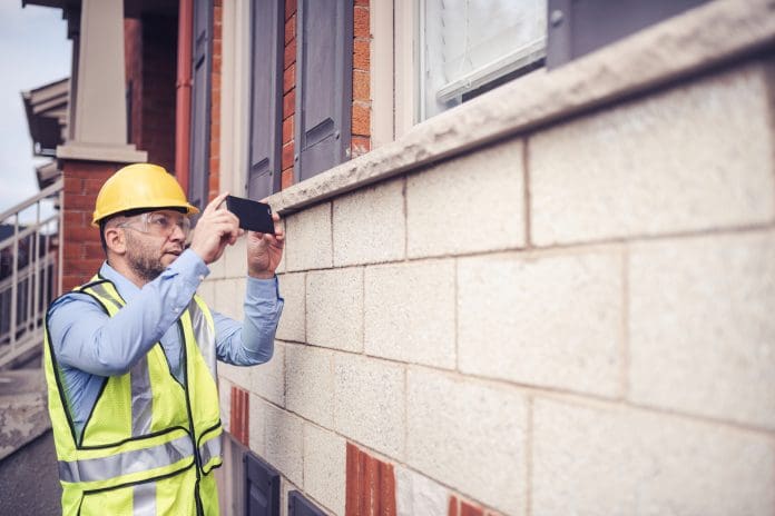 Building inspector, architect, engineer, general contractor, repairman insurance adjuster, or other blue collar worker inspecting the exterior walls of residential building. Wearing a safety vest, yellow helmet and clear safety glasses, he is checking very meticulously every deficiency and photographs them using his large screen phone, representing efficacy cover