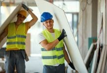 Construction Skills Mission Board to help recruit extra 100,000 workers by 2029 Two young Caucasian male construction workers are cheerfully carrying together pieces of drywall, representing young workers recruited by the Construction Skills Mission Board