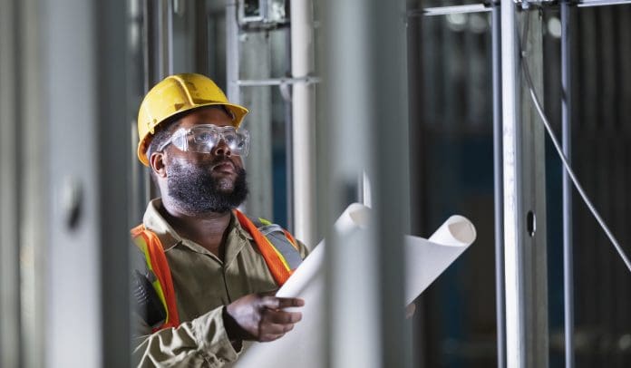 A mid adult African-American man working at a construction site wearing a yellow hardhat, holding a set of blueprints, and examining the structure being built. He is in his 30s, looks a bit confused and represents the ACQP's call for the removal of the Building Safety Regulator from the HSE