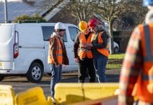 What to do after a commercial fleet accident: insurance and legal steps Wide-view of three construction workers engaged in a discussion at an outdoor worksite in Seghill, North East England. They're wearing safety gear, including hard hats and high-visibility vests, while standing together on site near construction barriers and their work van. One construction worker is using a digital tablet, representing what to do after a commercial fleet accident