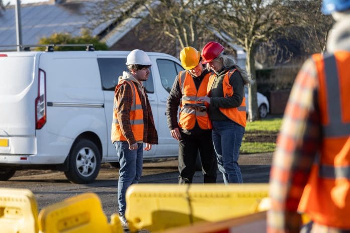 Wide-view of three construction workers engaged in a discussion at an outdoor worksite in Seghill, North East England. They're wearing safety gear, including hard hats and high-visibility vests, while standing together on site near construction barriers and their work van. One construction worker is using a digital tablet, representing what to do after a commercial fleet accident