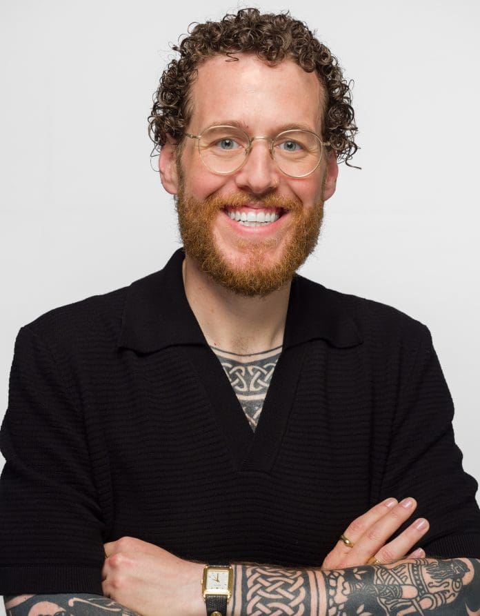 Jake Bush of RLB looking towards the camera against a white backdrop, smiling with arms folded