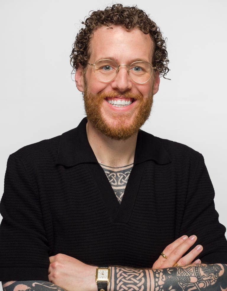 Jake Bush of RLB looking towards the camera against a white backdrop, smiling with arms folded