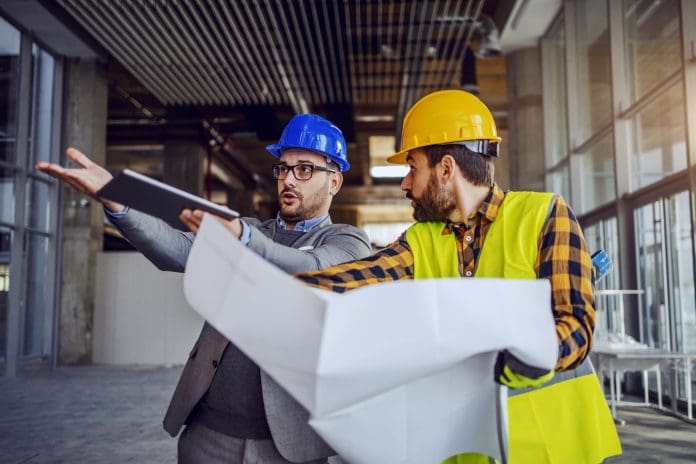 Upset architect arguing with construction worker and showing him his mistake. Worker holding blueprints and defending himself. Building in construction process interior, representing poor understanding of the Building Safety Act