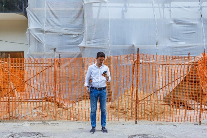 A young Caucasian man is standing in front of a construction site, using his phone and holding a file folder, representing the BSR backlog