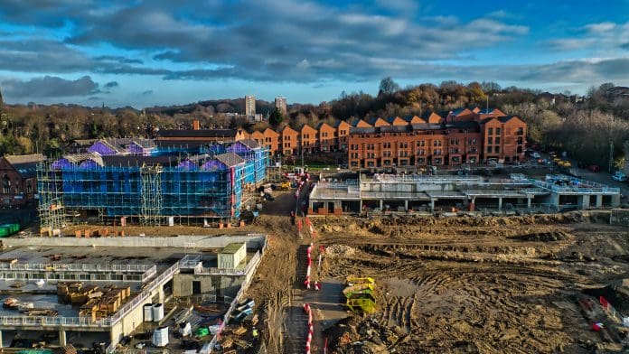 Aerial View of Construction Site with New Townhouses and Scaffolding in Kirkstall, Leeds. Aerial view of a construction site featuring a partially completed building wrapped in blue scaffolding, new brick townhouses, and extensive earthworks. The sky is partly cloudy.