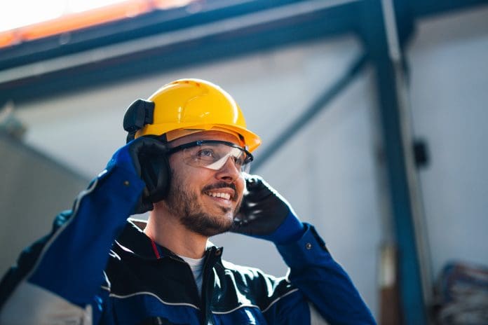 A cheerful male worker wearing a safety helmet and protective gear speaks on a headset in a bustling factory environment, emphasizing industry safety and productivity. It’s important to understand what factors to consider when selecting a warranty provider for your build projects