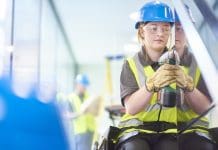 Building the future: Why apprenticeships are vital for Building Control a teenage female carpenter is installing handrails in a modern office refurbishment. IN the background a co-worker can be seen holding plans . They are wearing safety workwear. Representing building control apprenticeships
