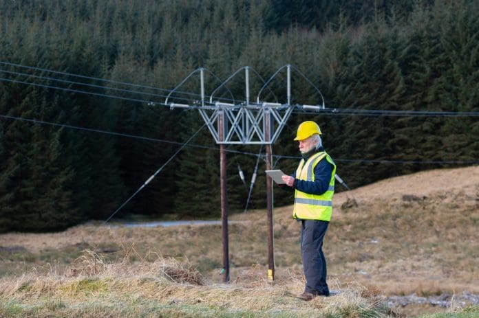 The rear view of a man wearing a high visibility waistcoat and hard hat using a digital tablet while standing close to high voltage power lines in rural Dumfries and Galloway south west Scotland, representing the SP Energy Networks framework