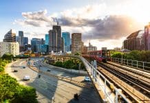 The UK has a 10-year Infrastructure Strategy: Now what? Elevated railroad tracks and passenger train with the modern skyline of the city of London in the distance. Below, a main road, with blurred motion of traffic, leads into the city, representing the 10 year infrastructure strategy