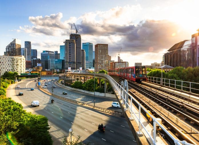 Elevated railroad tracks and passenger train with the modern skyline of the city of London in the distance. Below, a main road, with blurred motion of traffic, leads into the city, representing the 10 year infrastructure strategy