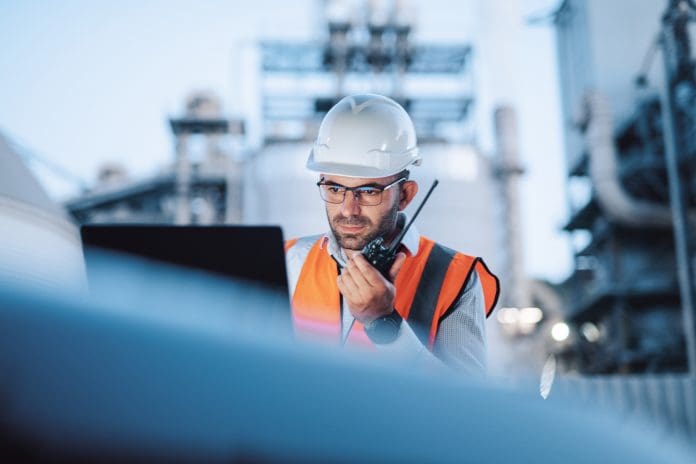 Why digital procurement platforms are powering construction’s digital leap in the age of AI A mid-adult male engineer works diligently during a night shift at an oil refinery, utilizing a laptop for monitoring operations and a walkie-talkie for real-time communication, representing digital procurement platforms