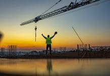 Certainty under pressure: Why confidence in construction matters more than ever Construction worker throwing up his hands in joy while standing on rooftop at sunset. Excited worker holding hard hat in his hand with arms up under the tower crane.