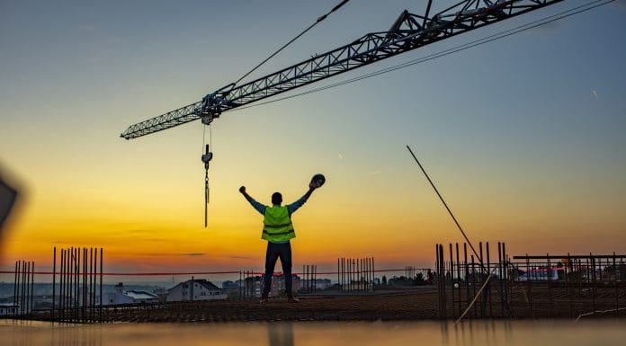 Construction worker throwing up his hands in joy while standing on rooftop at sunset. Excited worker holding hard hat in his hand with arms up under the tower crane.