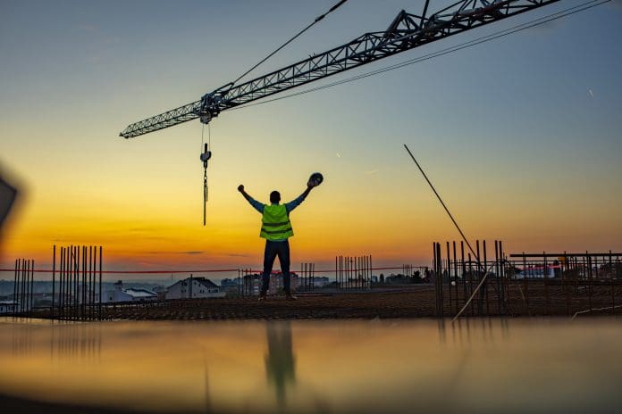 Success! Construction worker throwing up his hands in joy while standing on rooftop at sunset. Excited worker holding hard hat in his hand with arms up under the tower crane.