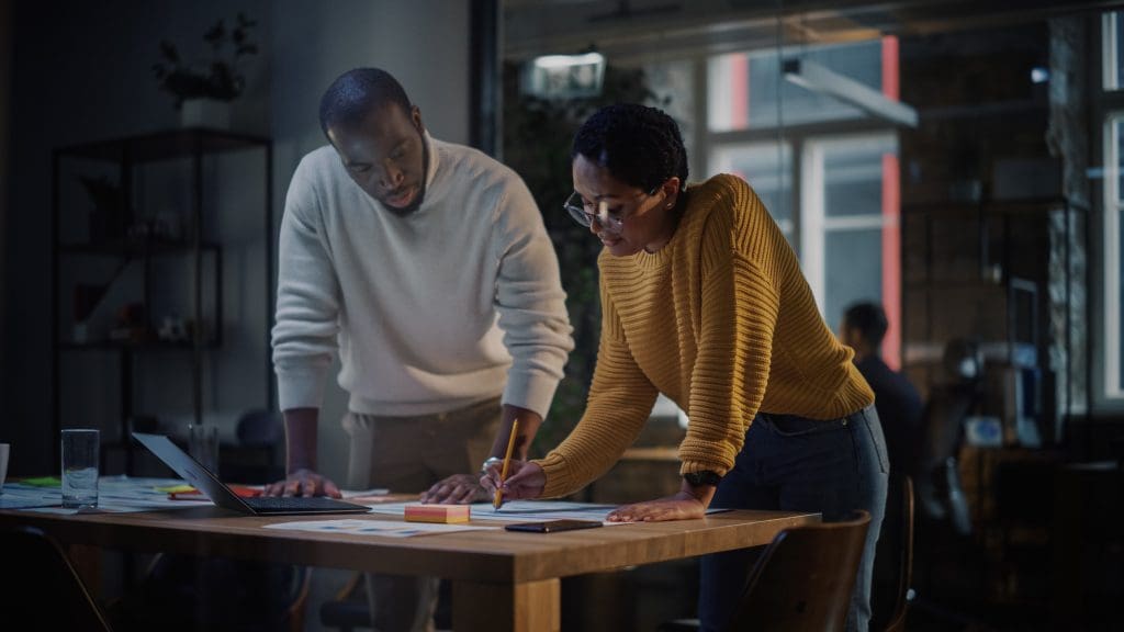 Two Diverse Multiethnic Colleagues Have a Conversation in a Meeting Room Behind Glass Walls in an Agency. African American Creative Director and Female Project Manager Discuss Work on Laptop Computer.