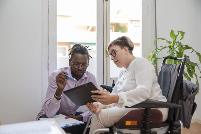 a businesswoman in a wheelchair with a disability looking at the digital tablet with her multiethnic business partner discussing the business plan, representing accessibility training