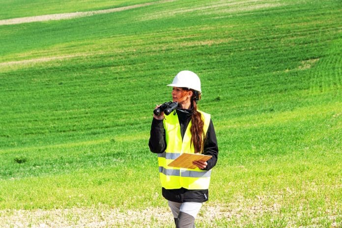 Female engineer wearing safety helmet and vest is walking on a green field, inspecting the area with binoculars and holding a clipboard, ensuring sustainable energy development, representing Land Quality Service