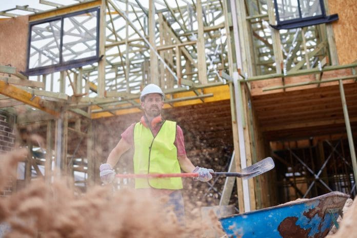 Worker throwing sand with shovel at site Male worker throwing sand with shovel against incomplete building. Confident engineer is working at construction site. He is in reflective clothing.