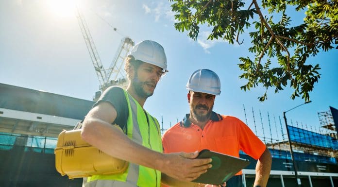 Two engineers wearing hardhats and high-visibility vests are discussing building plans at a construction site in australia, representing Project Management Day