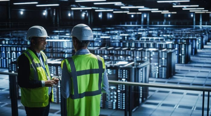 Data Center IT Specialist and System administrator Talk, Use Tablet Computer, Wearing Safety Wests. Server Clod Farm with Two Information Technology Engineers checking Cyber Security.