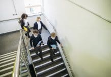 DfE confirms cuts to free schools building programme High angle view of British teenagers in uniforms carrying backpacks and notebooks as they move down steps in education building, representing Free schools building programme cuts