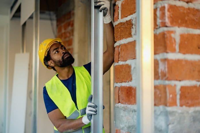 Worker in protective gear and safety vest inspecting a metal frame during construction. Bright lighting highlights tools, showcasing work in progress for a building interior concept, representing material passports