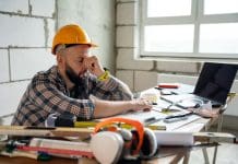 Mental health: The hidden cost of late construction payments An exhausted worker wearing a safety helmet, leaning on drawn blueprints amidst construction tools and materials at a desk, illustrating fatigue and effort in architectural or construction projects. Representing the impact of late construction payments