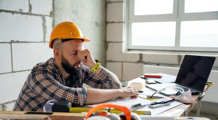 An exhausted worker wearing a safety helmet, leaning on drawn blueprints amidst construction tools and materials at a desk, illustrating fatigue and effort in architectural or construction projects. Representing the impact of late construction payments