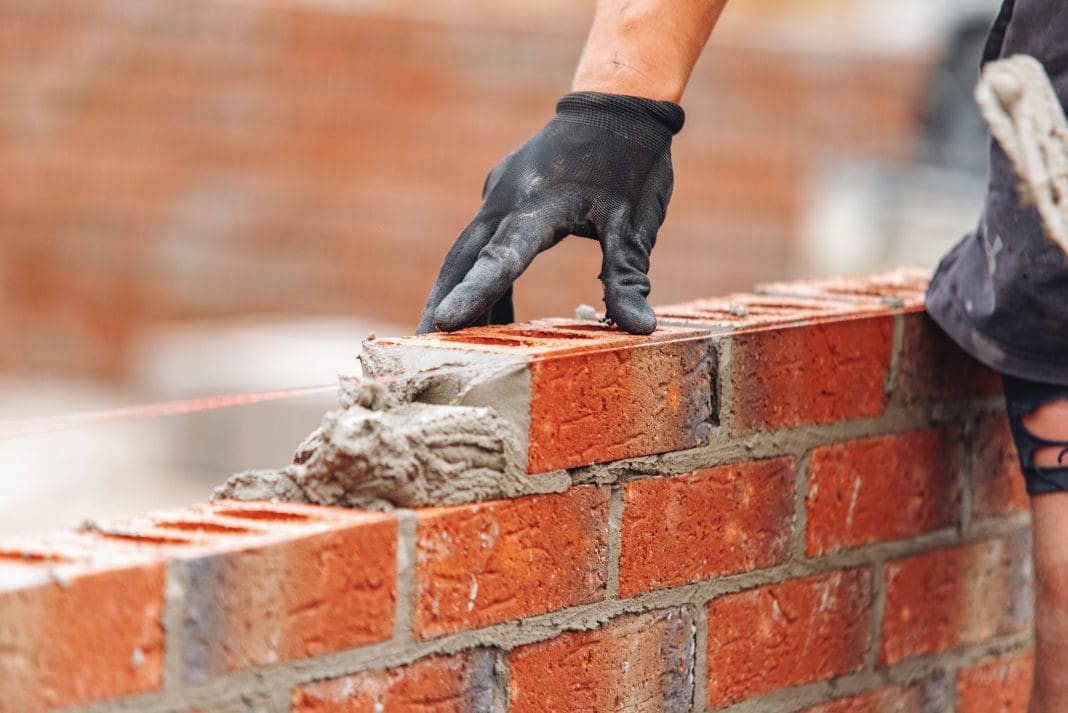 Close-up of Bricklayer constructing brick wall using red bricks, cement and tools while wearing gloves, representing falling construction output