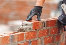 Latest ONS figures see construction output fall by 0.3% Close-up of Bricklayer constructing brick wall using red bricks, cement and tools while wearing gloves, representing falling construction output
