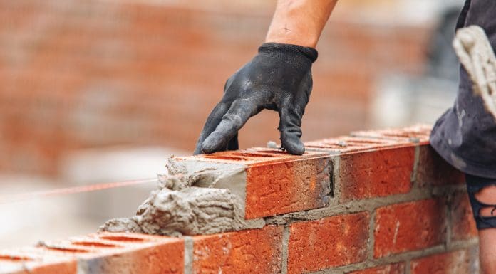 Close-up of Bricklayer constructing brick wall using red bricks, cement and tools while wearing gloves, representing falling construction output