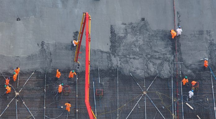 An aerial view captures construction workers in bright orange safety vests and hard hats spread across a foundation site as they coordinate concrete pouring from a red concrete pump truck with its extended boom, spreading and leveling the fresh wet concrete across geometric steel rebar reinforcement grids laid over the dark foundation surface, representing the top stories on PBC Today in 2025