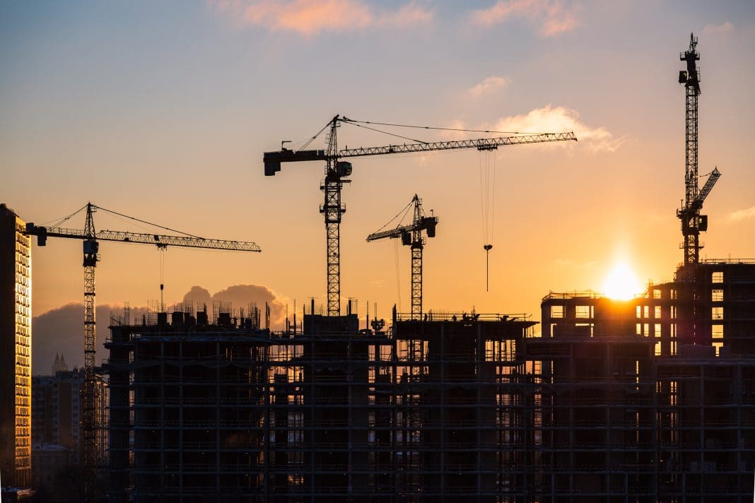 Lots of tower cranes build high-rise residential building in evening time. Steel frame structure in sunlight. Industrial background with sun flare. Yellow sunset sky, representing construction new orders