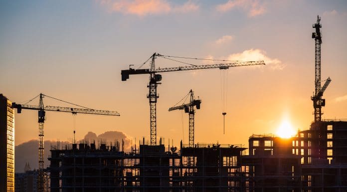 Lots of tower cranes build high-rise residential building in evening time. Steel frame structure in sunlight. Industrial background with sun flare. Yellow sunset sky, representing construction new orders