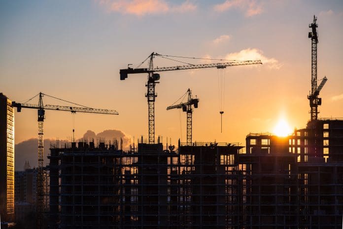Mace Consult Market View reports surge in new orders in construction Lots of tower cranes build high-rise residential building in evening time. Steel frame structure in sunlight. Industrial background with sun flare. Yellow sunset sky, representing construction new orders