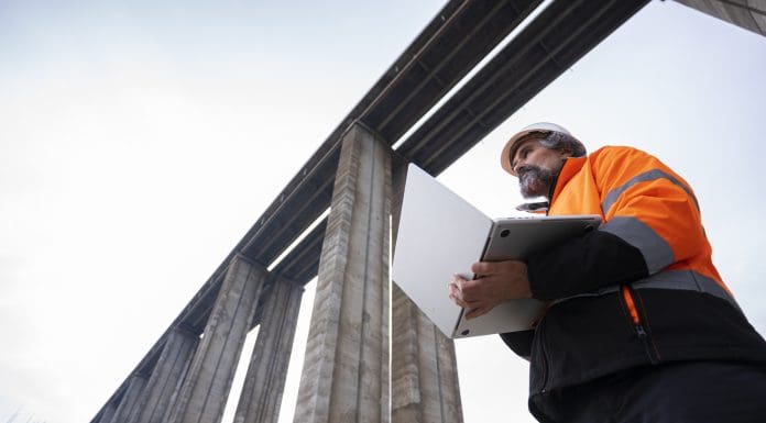 Experienced road engineer in safety gear & work helmet controls viaduct highway construction project using AI software on laptop. Building smart infrastructures