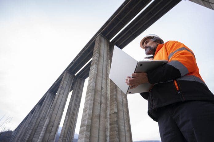Building smarter: Five IT trends set to shape construction in 2026 Experienced road engineer in safety gear & work helmet controls viaduct highway construction project using AI software on laptop. Building smart infrastructures