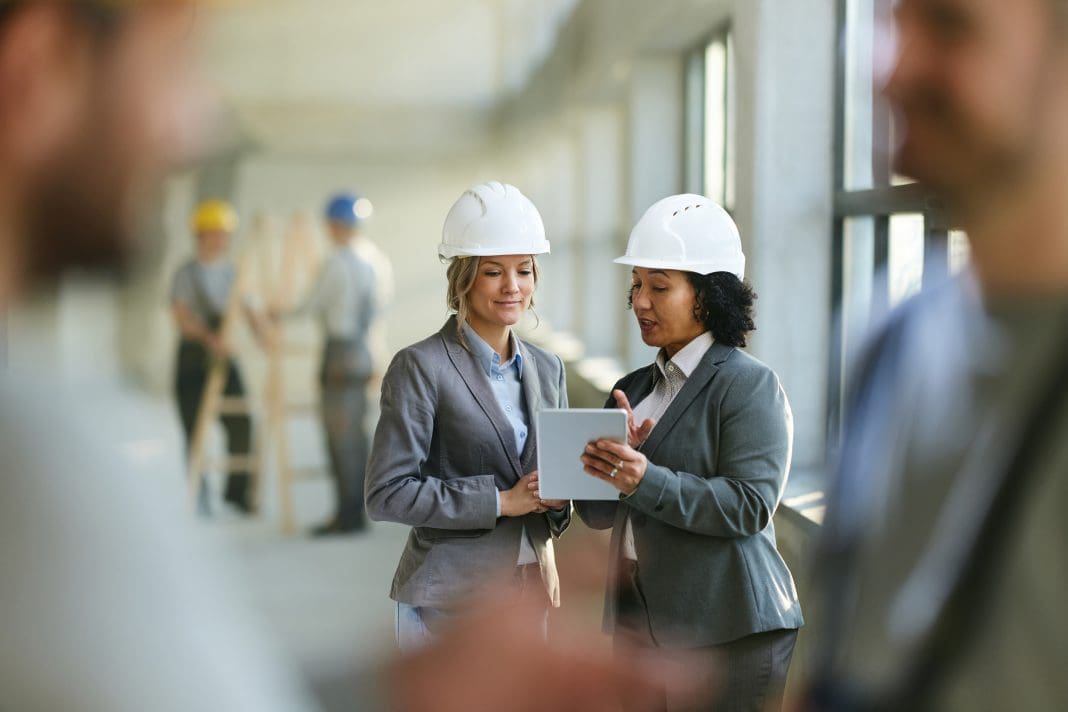 Female real estate developers cooperating while surfing the Internet on digital tablet at construction site, representing construction email management