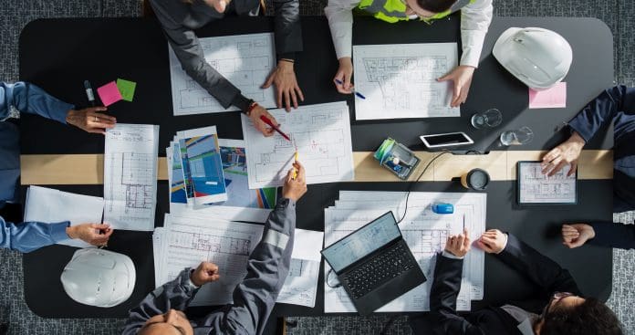 University of Cambridge joins CCBP Academic Advisory Group Top Down View of Diverse Team of Business Professionals and Engineers Working Together on Architectural Plans in a Corporate Office. Focused Colleagues Using Laptop, Tablet, Blueprints and Documents.