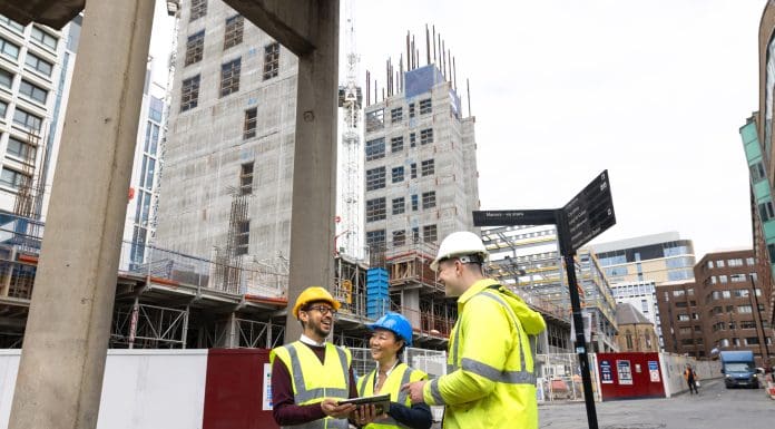 A wide shot of three construction employees standing together outside a construction site. They are discussing plans together using blueprints and an electronic tablet. The two men and woman are wearing high-visibility clothing and hard hats, representing a new personal professional experience consultation
