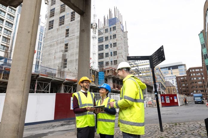 A wide shot of three construction employees standing together outside a construction site. They are discussing plans together using blueprints and an electronic tablet. The two men and woman are wearing high-visibility clothing and hard hats, representing a new personal professional experience consultation