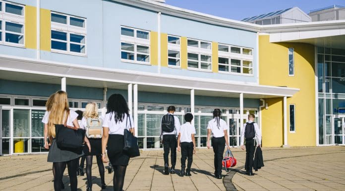 Rear view of male and female students walking towards school entrance on way in to school, punctuality, good behaviour, order. School children walking to school, representing Technical Advisor Services 2025 framework