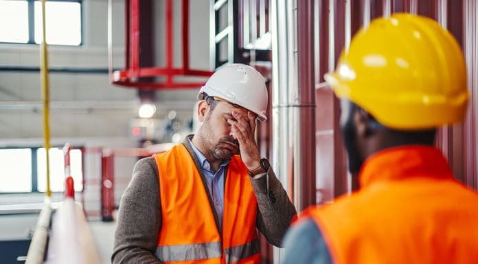 Adult engineer feeling overwhelmed and having a headache, covering his face with his hand while standing in a factory during a difficult work situation, representing construction critical distress