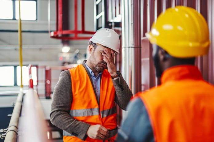 Adult engineer feeling overwhelmed and having a headache, covering his face with his hand while standing in a factory during a difficult work situation, representing construction critical distress